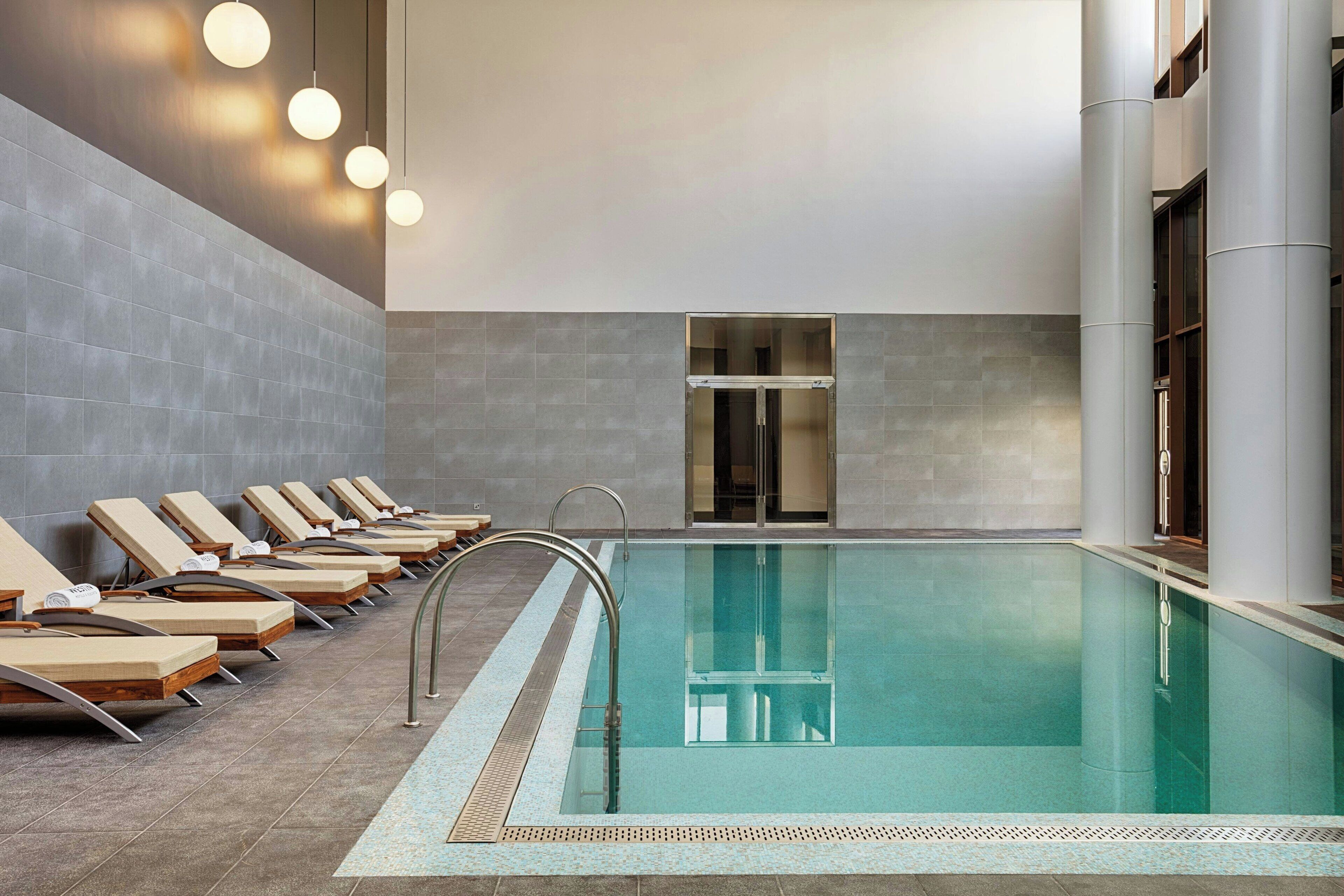Una piscina interior moderna con sillones de descanso y un muro de cristal.