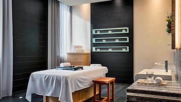 A modern hotel room with a bed, a wooden nightstand, a small table, a wall-mounted shelf with decorative items, and a bathroom with a sink and mirror.
