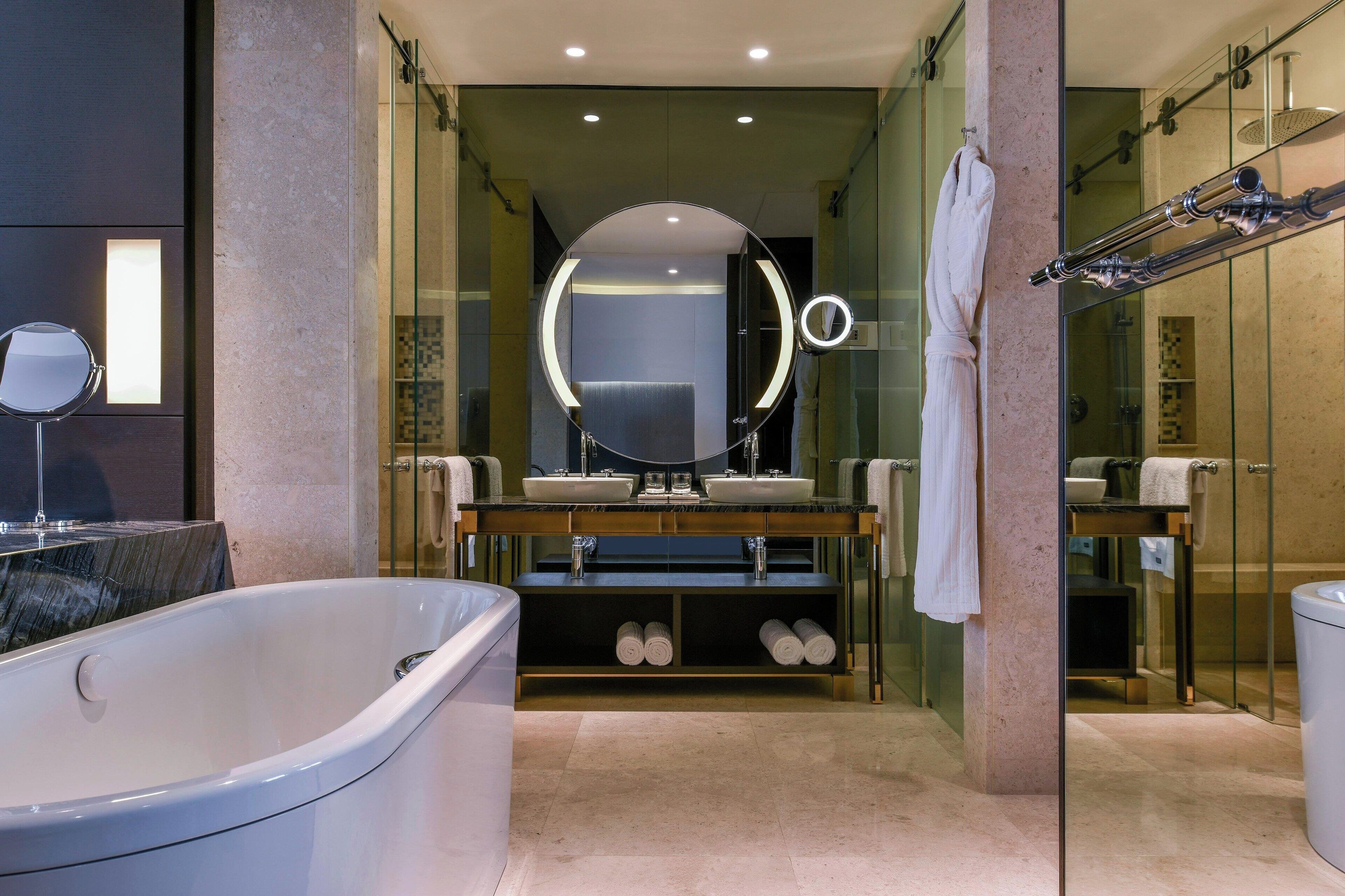 A modern bathroom with a large bathtub, dual sinks, a round mirror with lights, and a glass shower enclosure.