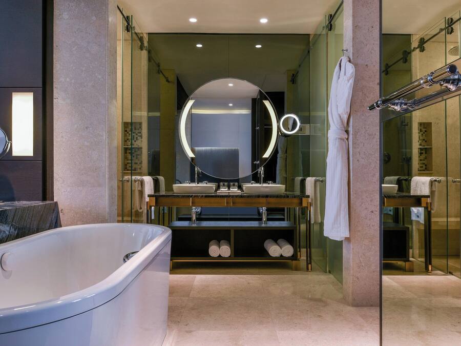 Un baño moderno con una bañera grande, lavamanos dobles, un espejo redondo con luces y un box de ducha de vidrio.
