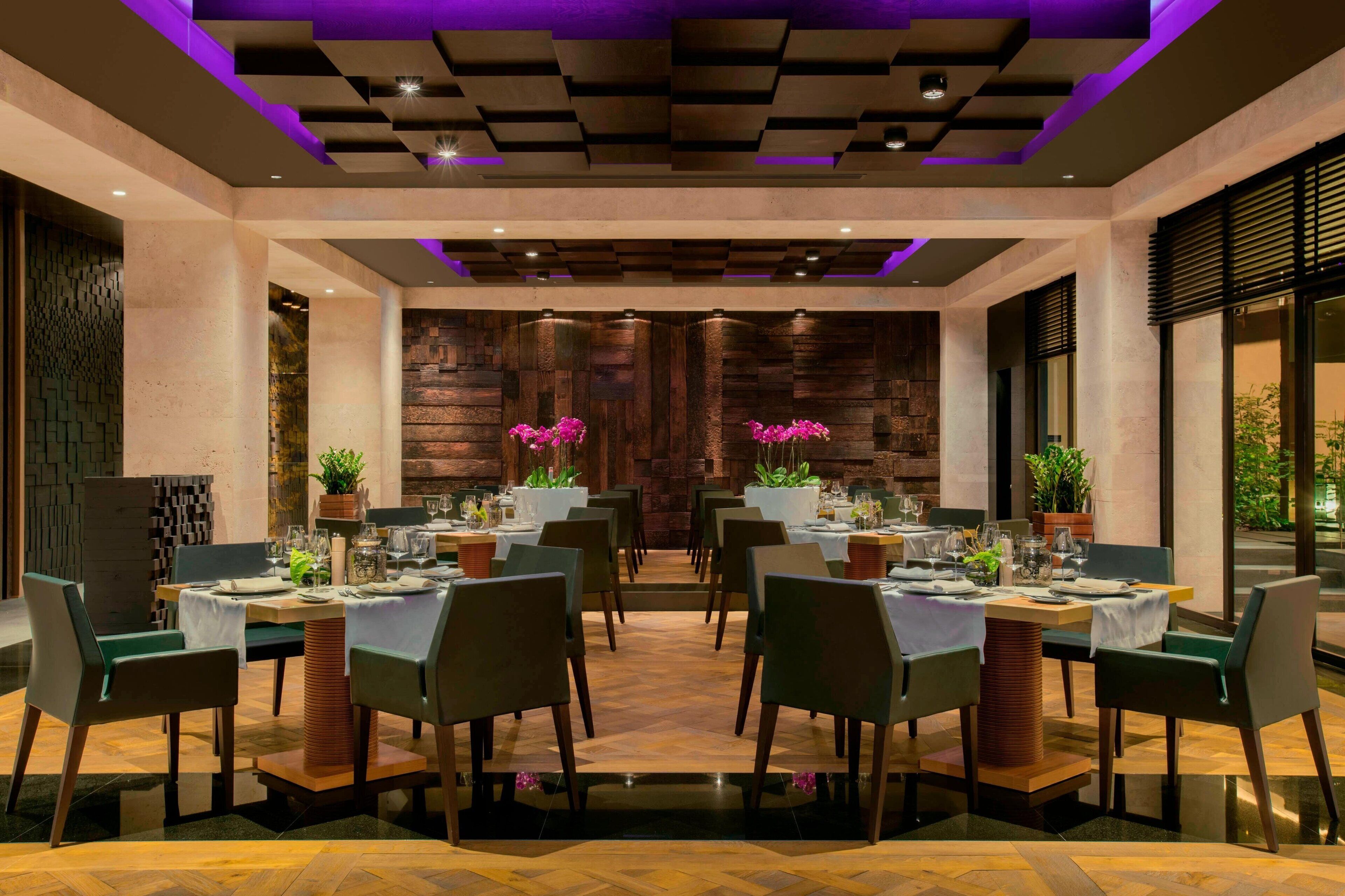 A restaurant with a central dining area featuring round tables, chairs, and potted plants.