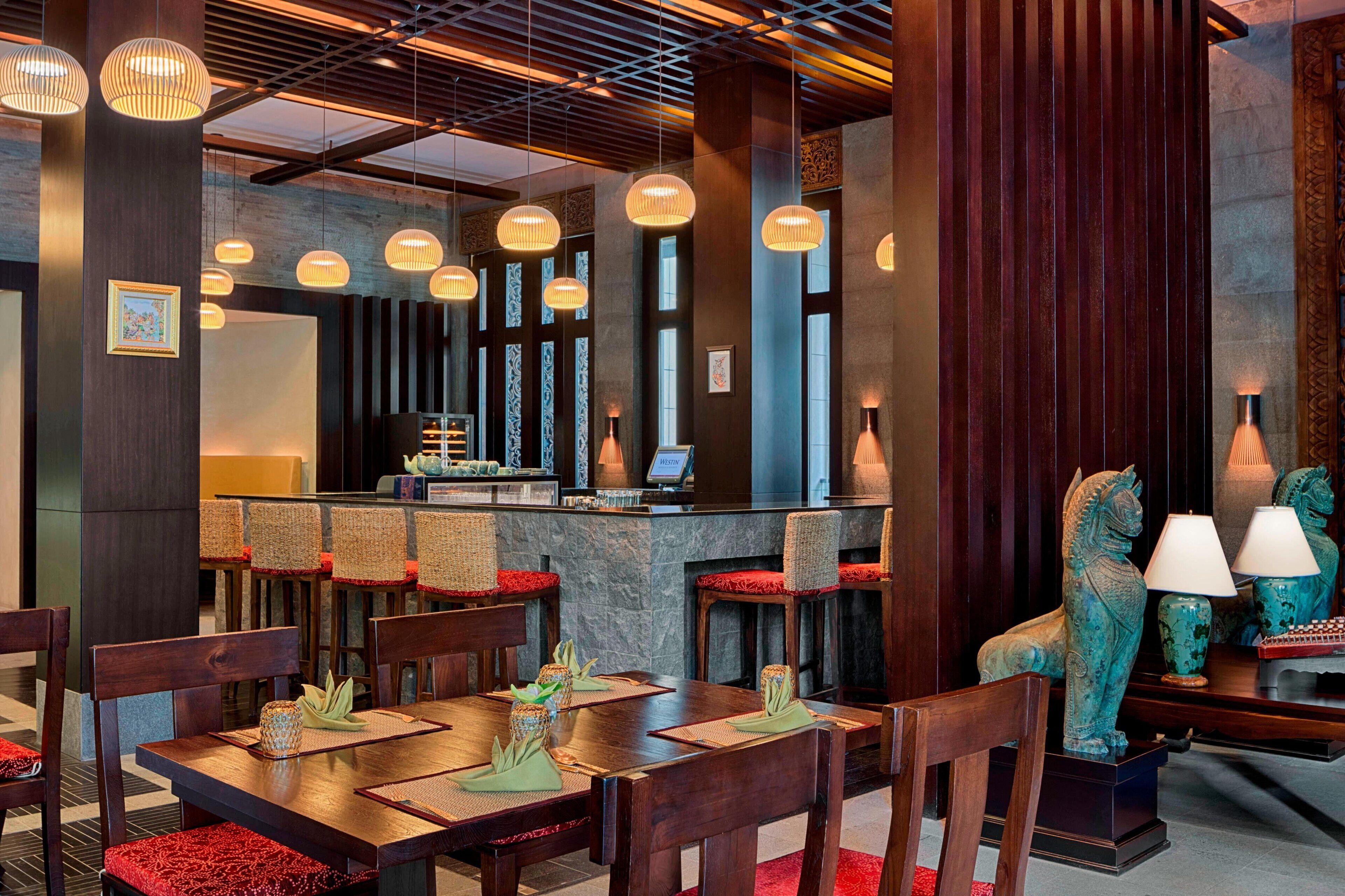 Un restaurant avec des tables et des chaises en bois, des lampes suspendues et des éléments décoratifs, comme un vase et une statue.