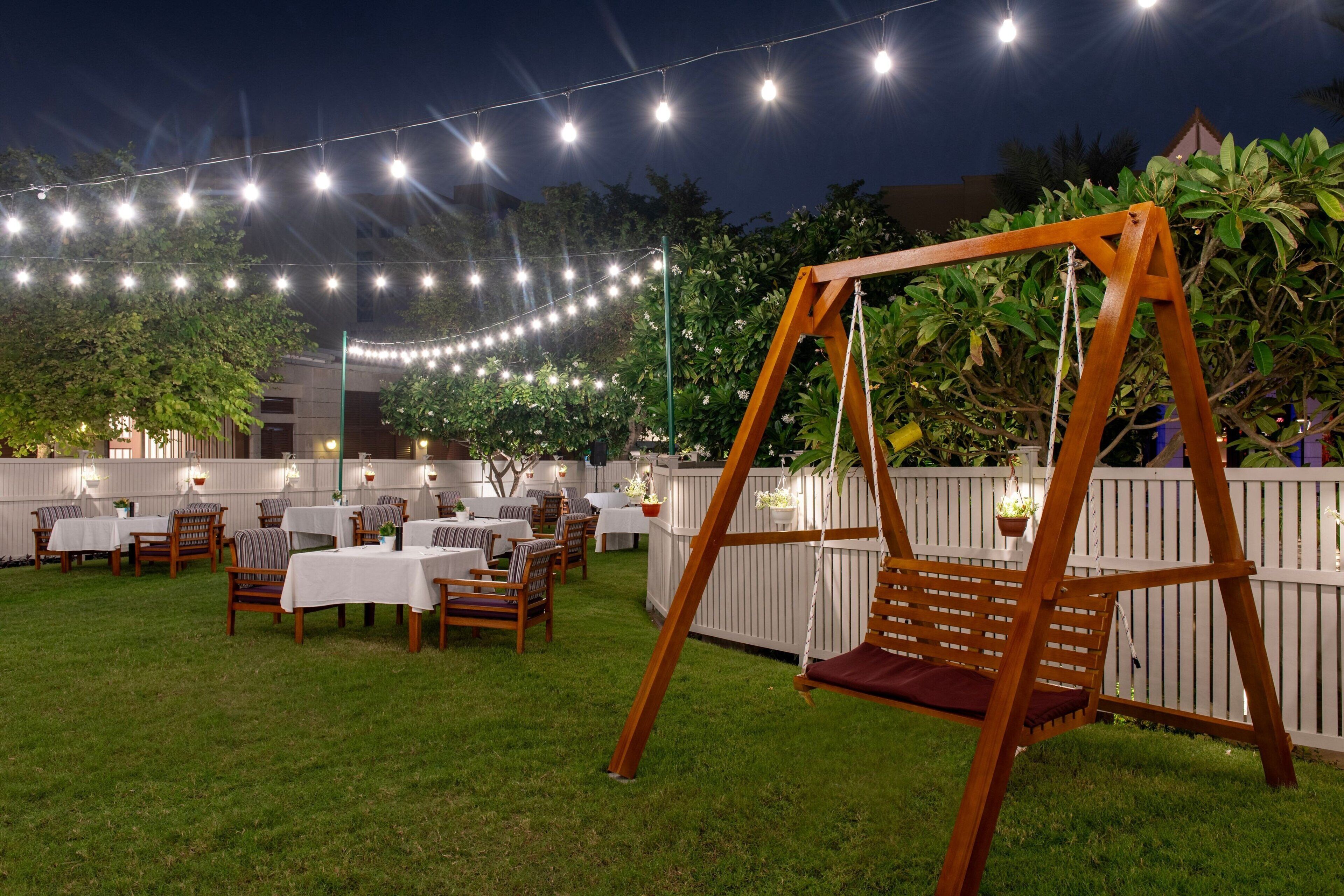 Un espace extérieur aménagé pour les repas, avec des guirlandes lumineuses, une balançoire en bois et des tables avec des chaises.