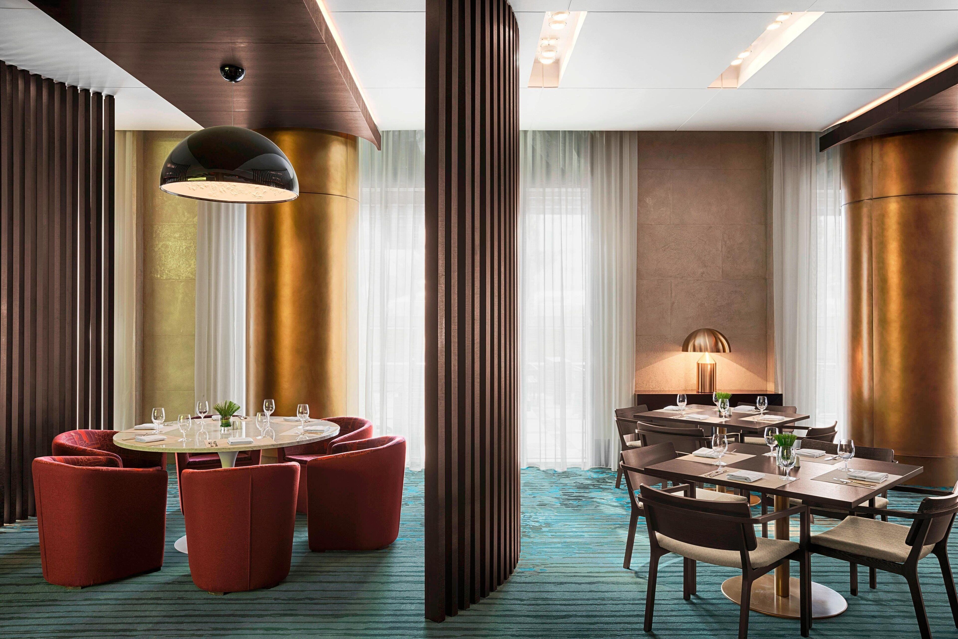 A modern hotel dining area with round tables, red upholstered chairs, and a large pendant light.