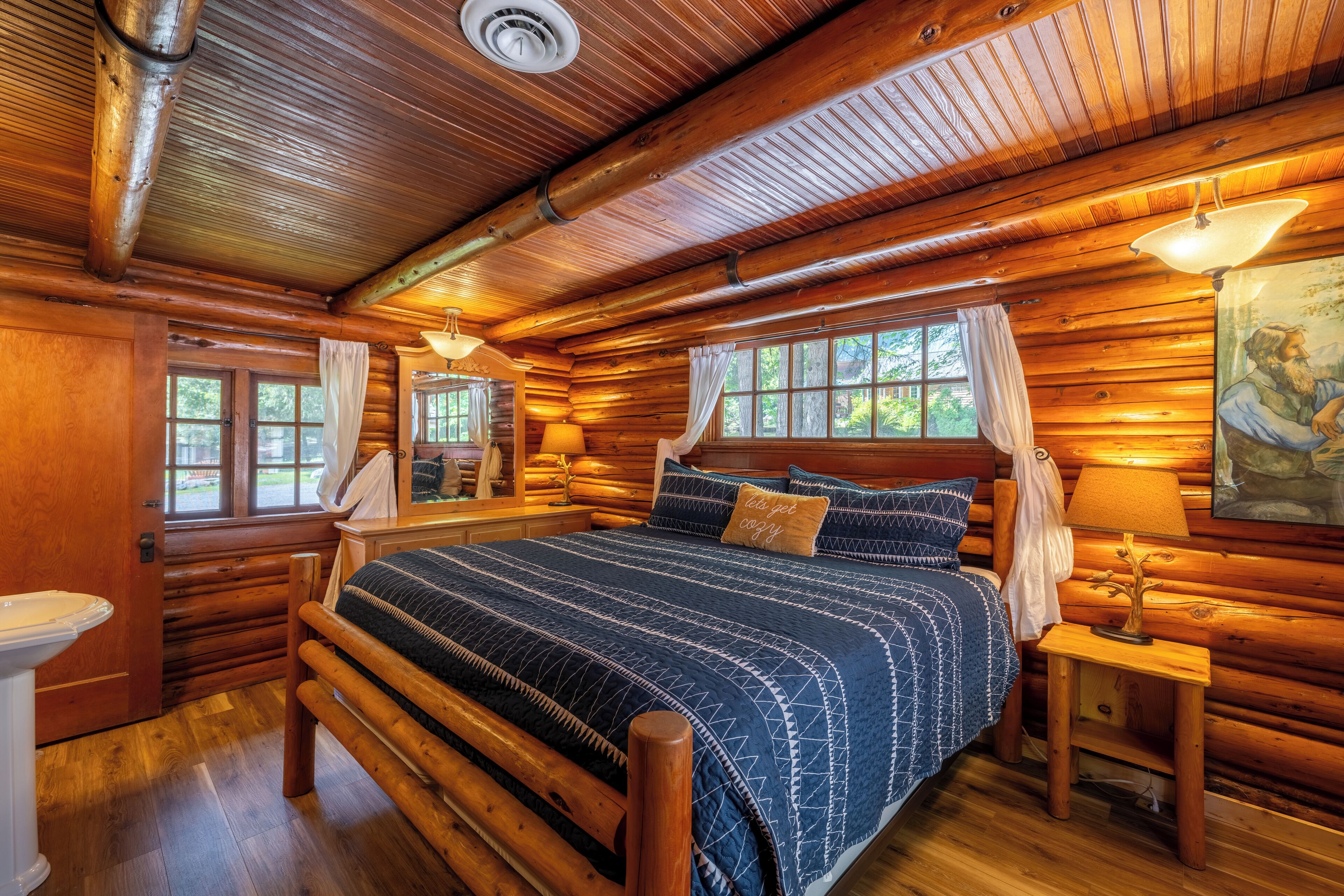 Ein Schlafzimmer in einer Blockhütte mit einem hölzernen Bett, einem Nachttisch, einem Fenster mit Vorhängen und einem Bild an der Wand.