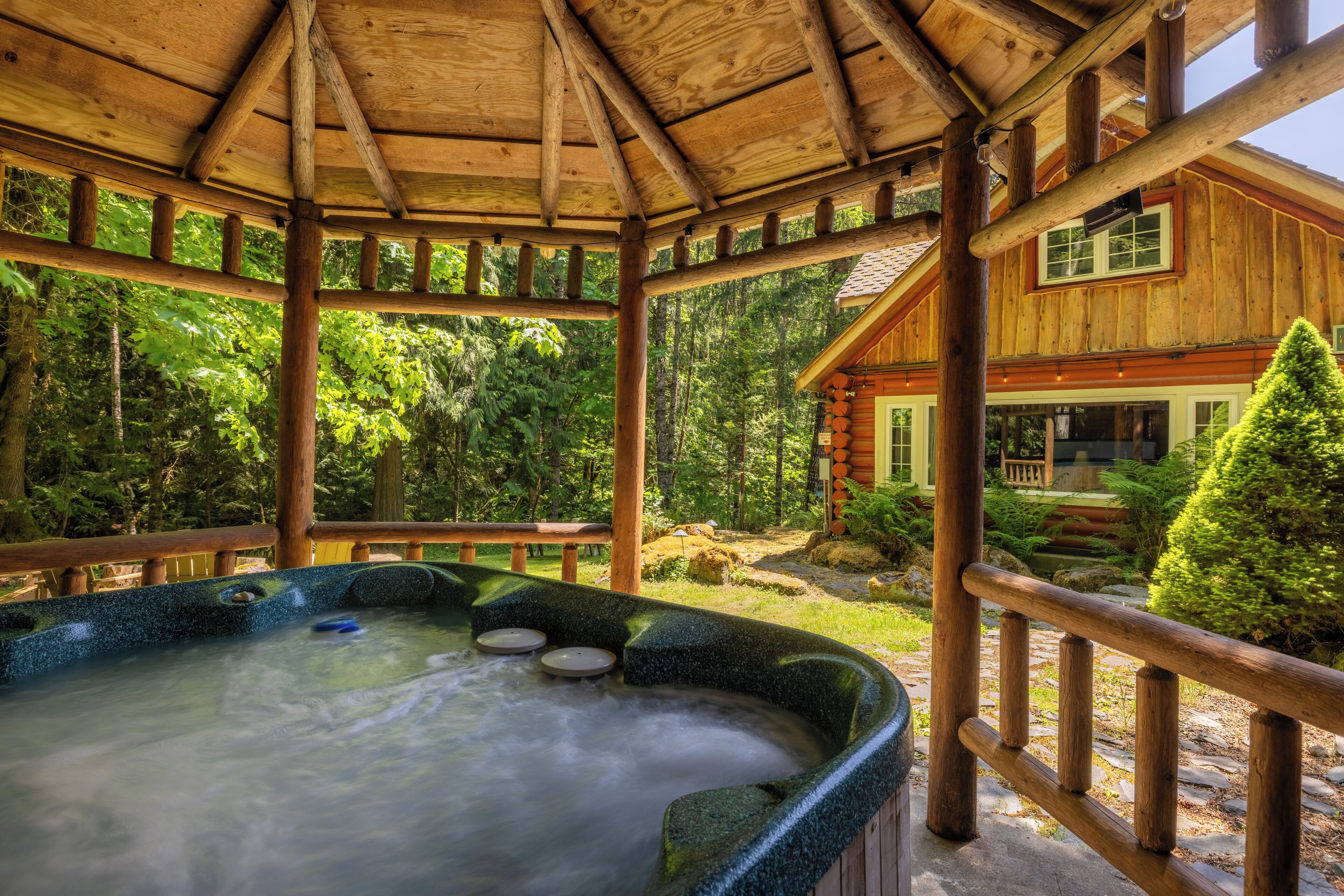 A wooden gazebo with a hot tub, surrounded by lush greenery and a wooden cabin.