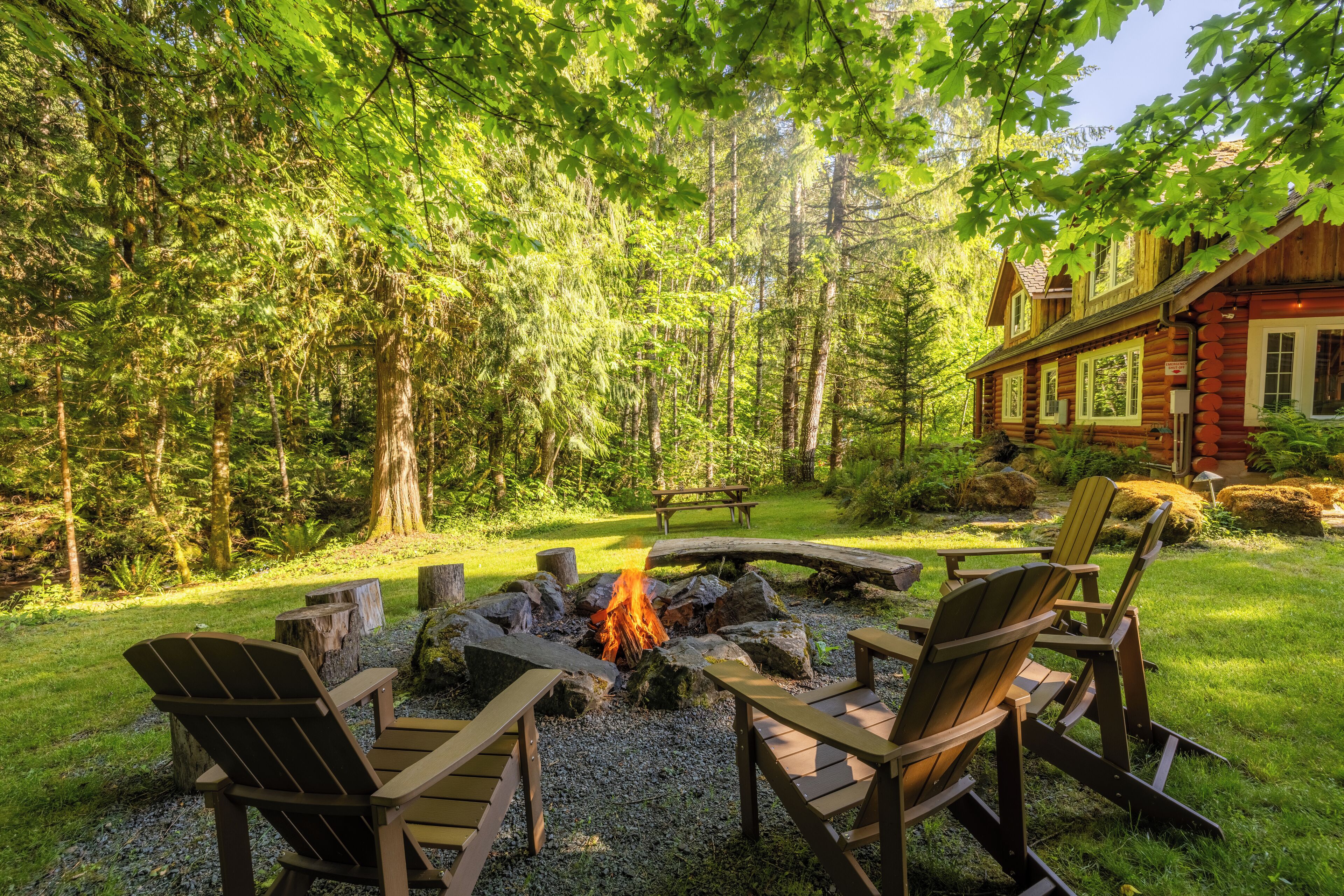 A log cabin with a fire pit surrounded by Adirondack chairs in a forest setting.