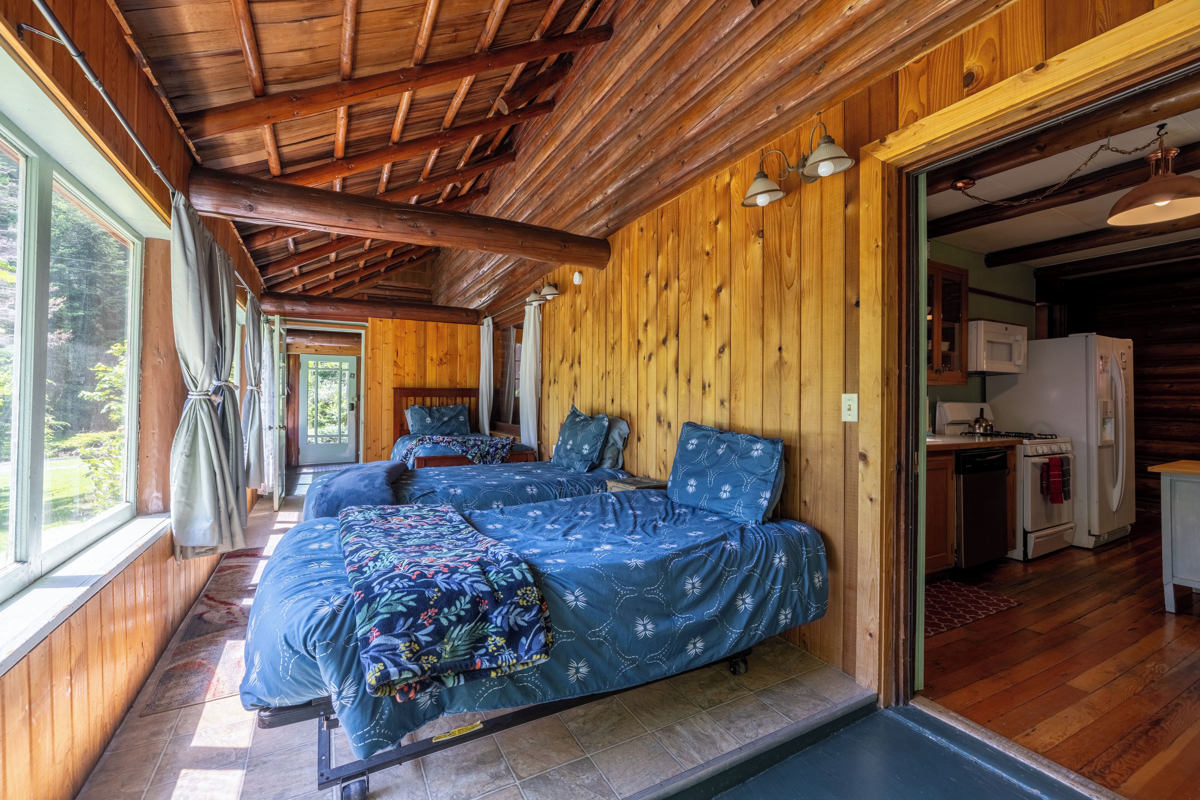 A cabin with two beds, wooden walls, a kitchen area, and large windows.