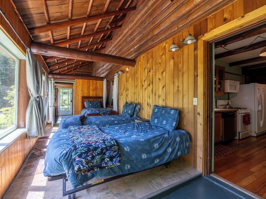 A cabin with two beds, wooden walls, a kitchen area, and large windows.