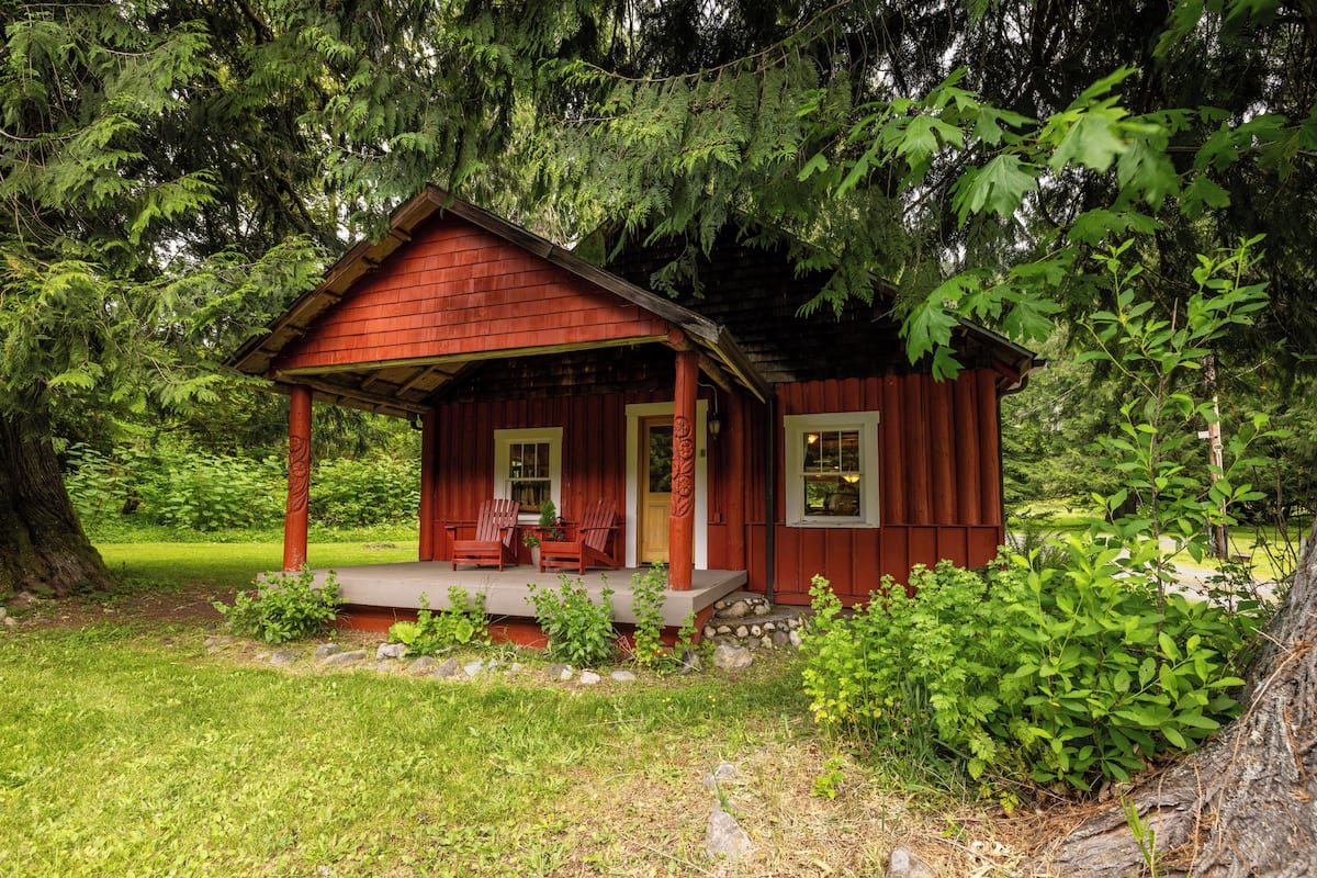 A small, rustic cabin with a porch and two chairs, surrounded by lush greenery.