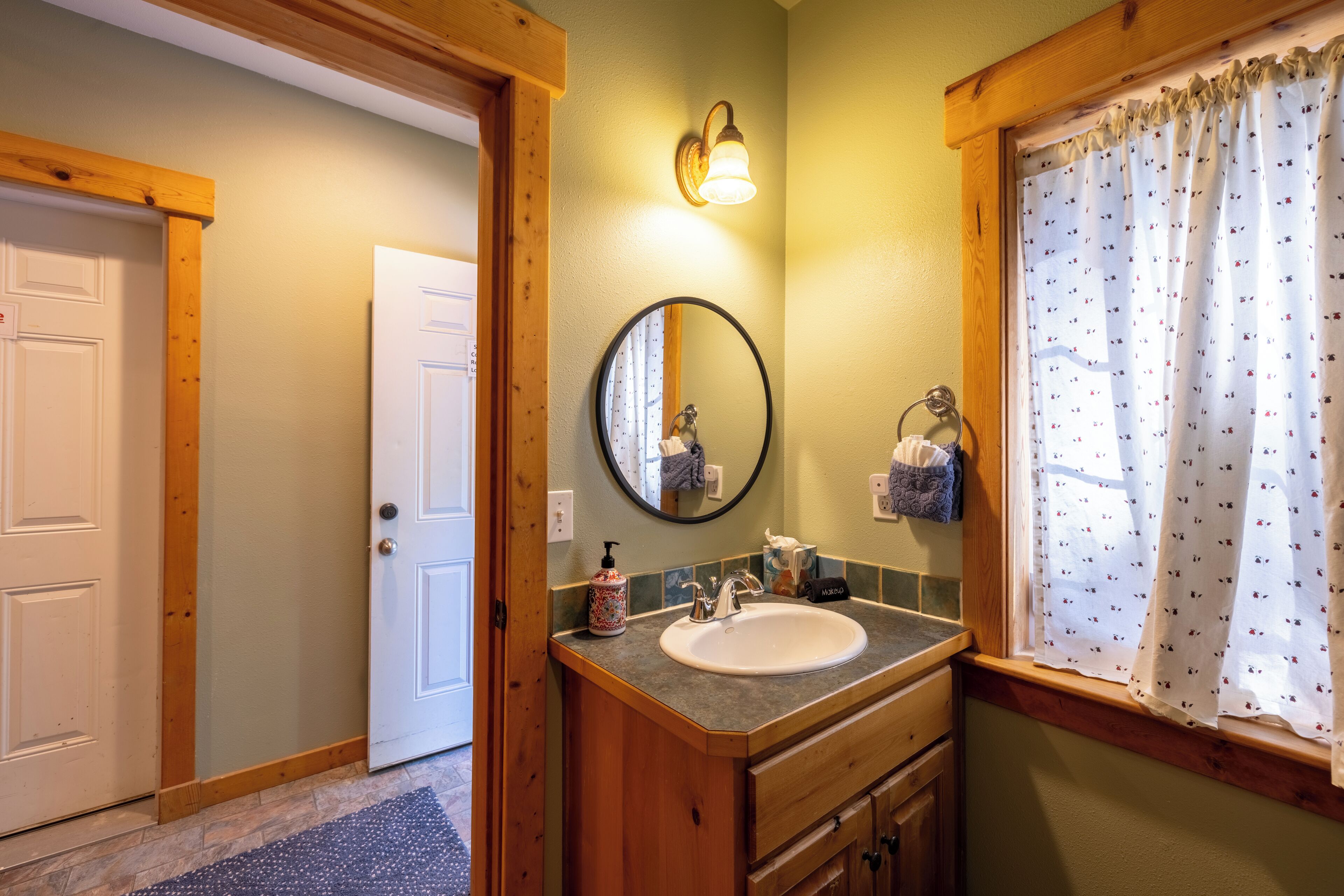 A bathroom with a wooden vanity, a round mirror, a sink, and a window with patterned curtains.
