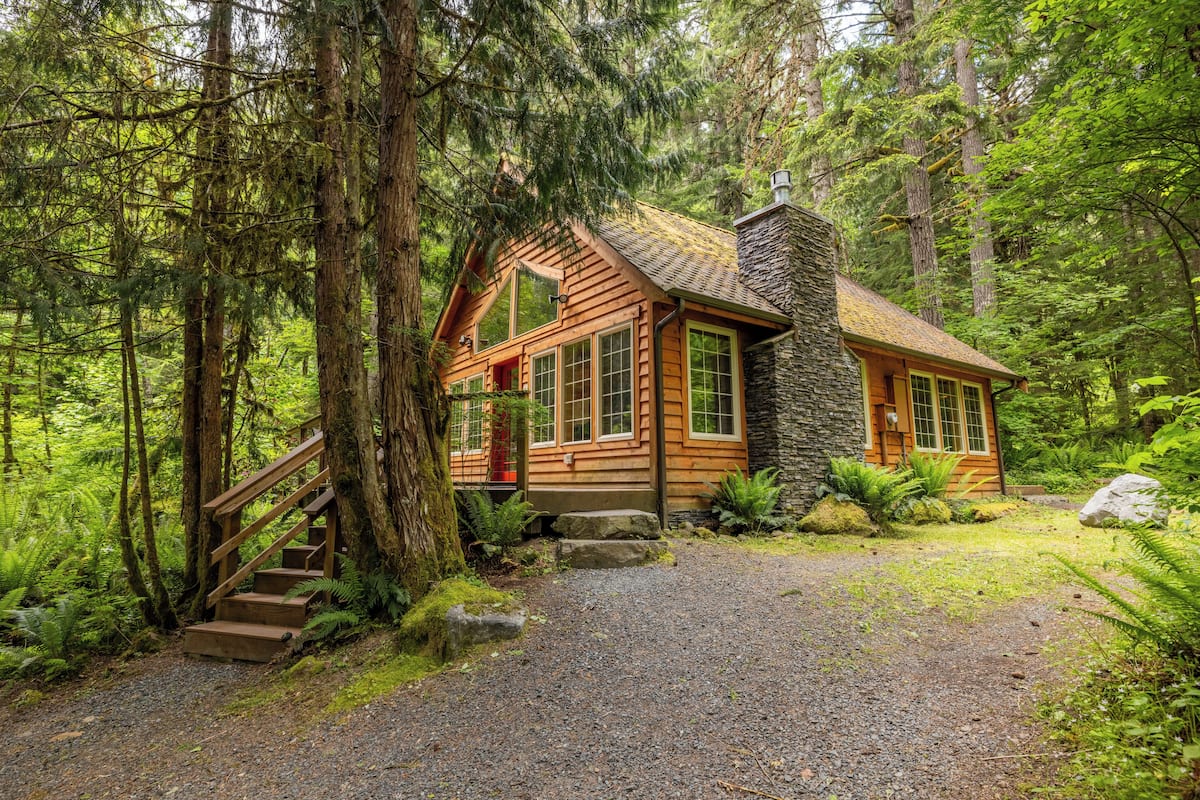 A rustic cabin with a stone chimney, wooden exterior, and a gravel path leading to a wooden staircase.