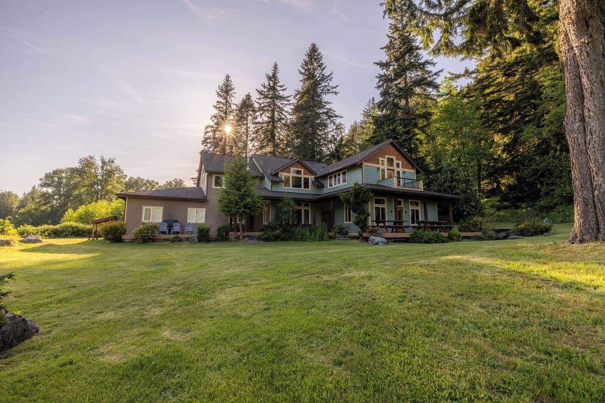 A large house with multiple sections, surrounded by a well-maintained lawn and tall trees.