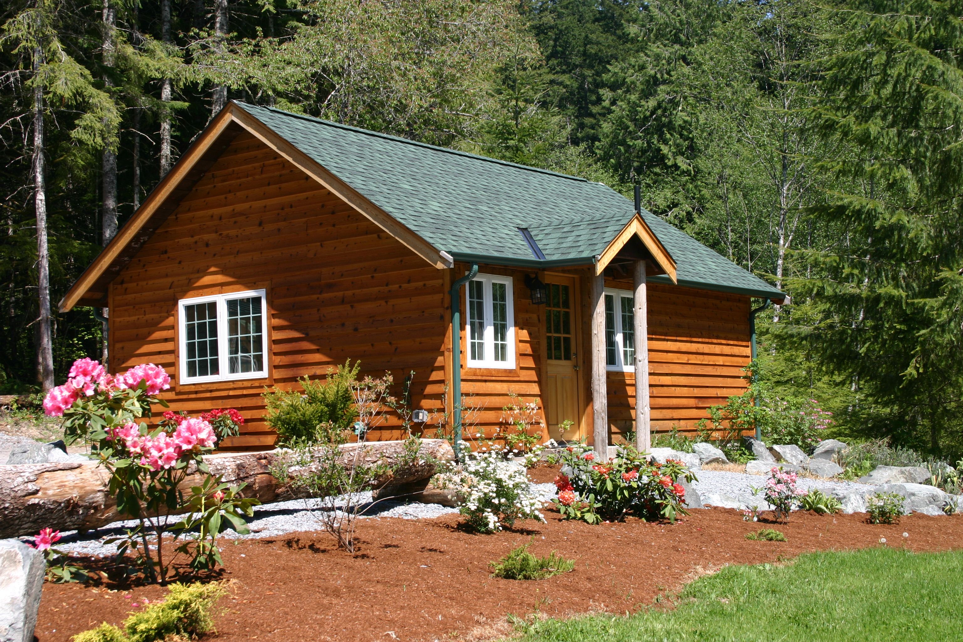 Uma cabana de toras com telhado verde, rodeada por um jardim com diversas plantas e flores.