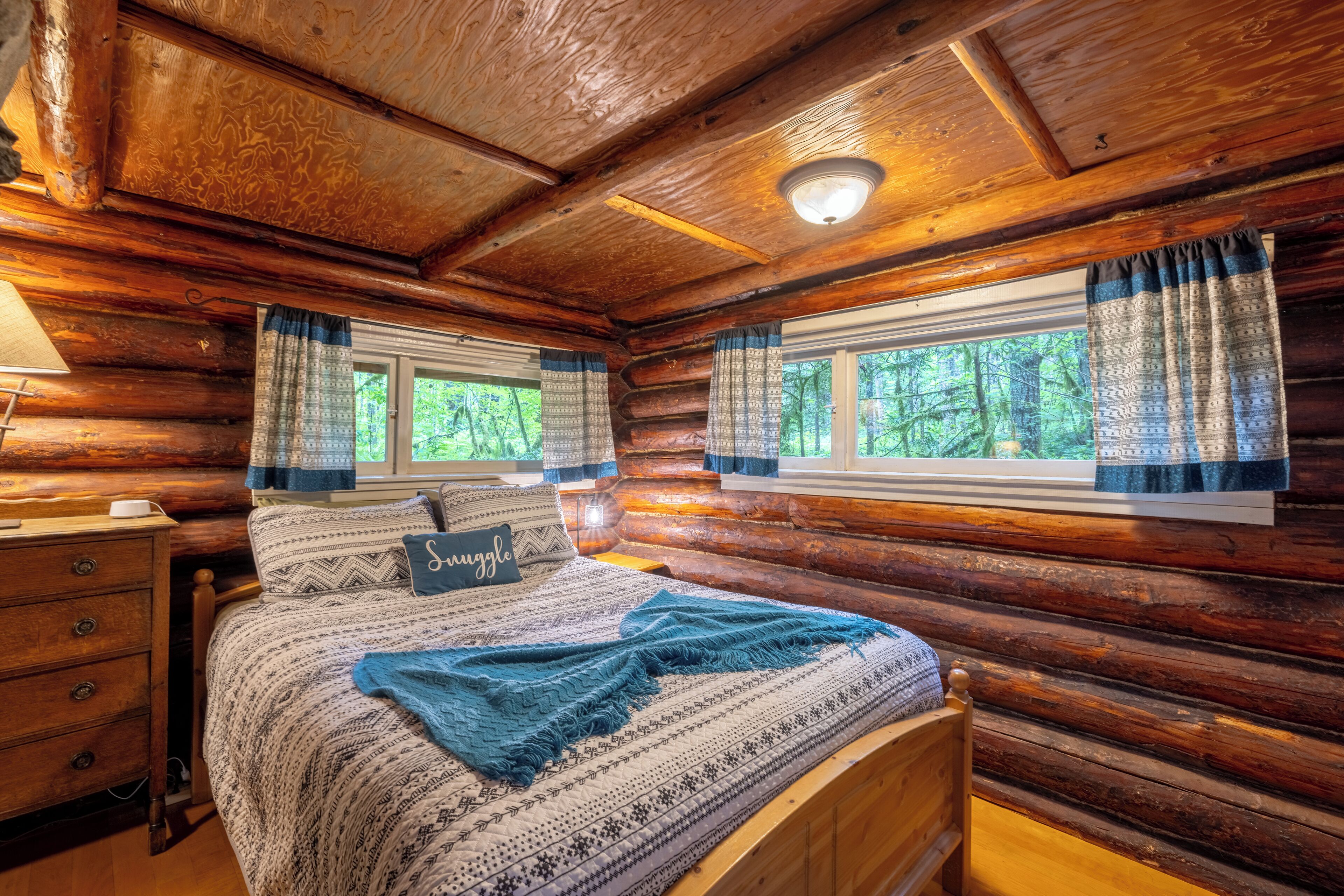 Quarto de cabana de tronco com cama, mesa de cabeceira e janela com cortinas.