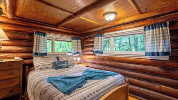 Quarto de cabana de tronco com cama, mesa de cabeceira e janela com cortinas.