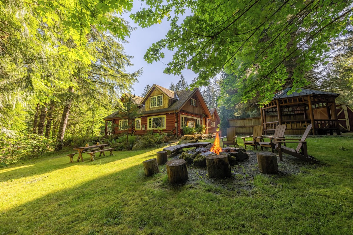 A cabin with a fire pit surrounded by wooden stumps, a picnic table, and a wooden deck.
