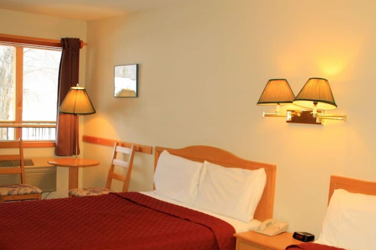 Quarto de hotel com duas camas, mesa de cabeceira, luminária e janela com cortinas.
