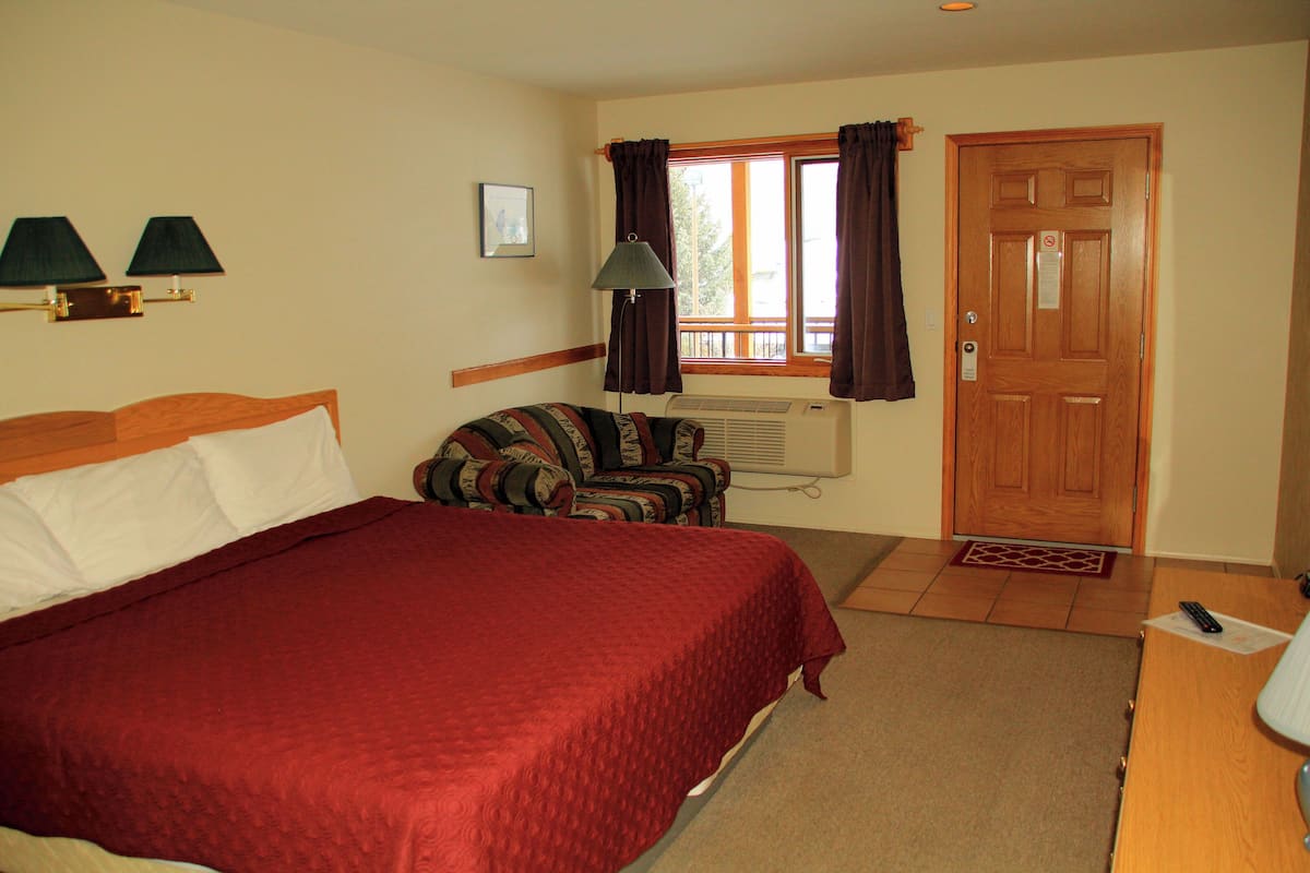 Quarto de hotel com uma cama grande, uma cadeira, uma escrivaninha, uma janela com cortinas, uma porta e uma luminária fixada na parede.
