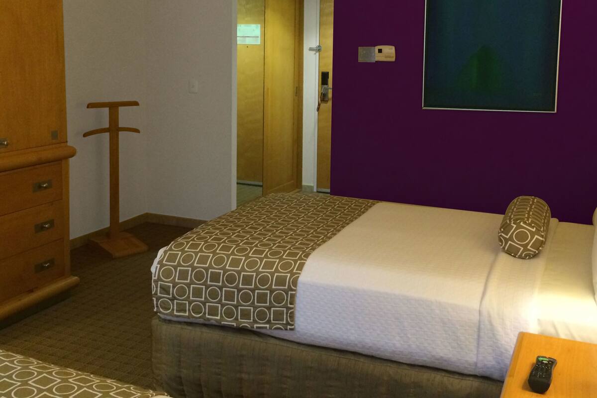 A hotel room with a bed, a dresser, a wooden stool, a remote control, and a purple wall.