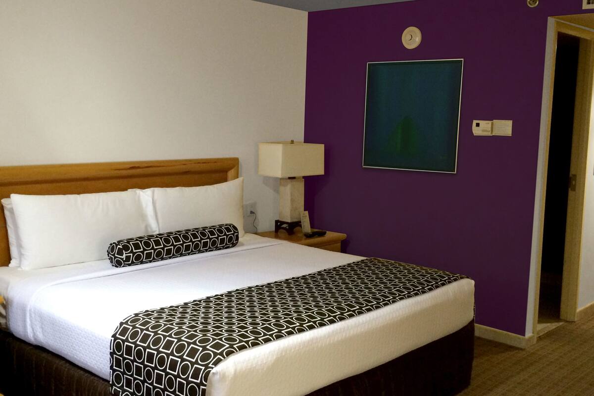 A hotel room with a bed, a nightstand, a lamp, a painting on the wall, and a door.