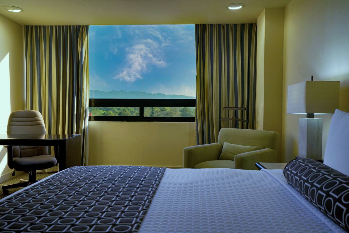 A hotel room with a large bed, two armchairs, a desk, and a view of mountains through a window.