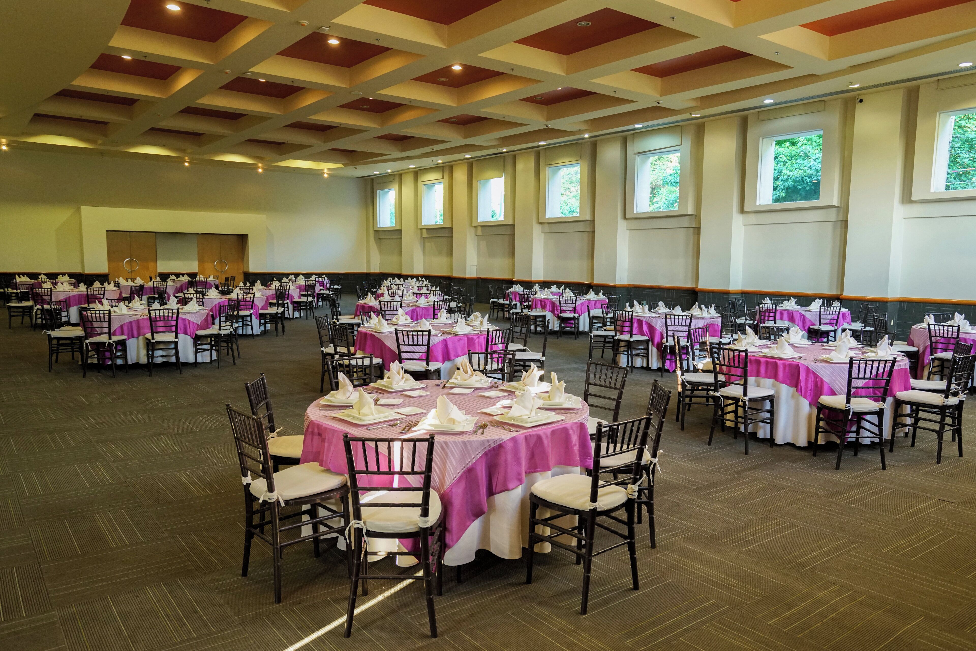 A large banquet hall with round tables set for events, featuring a high ceiling with wooden beams and large windows.