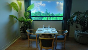 A dining area with a table set for four, surrounded by chairs, with large potted plants on either side and a window with a view of greenery. - Gamma by Fiesta Americana Xalapa Nubara (Xalapa)