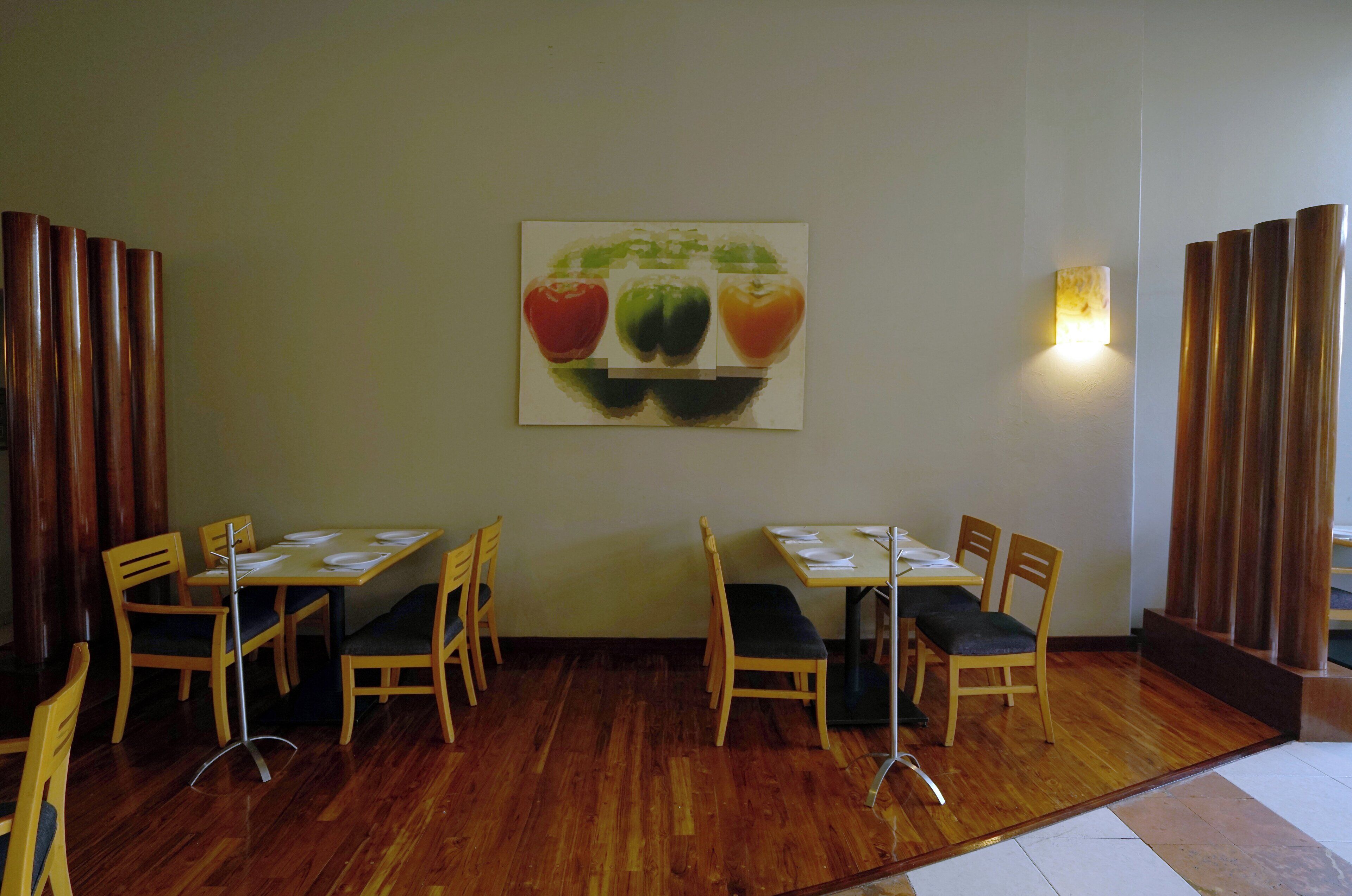 A dining area with wooden tables and chairs, a wall-mounted painting of bell peppers, and a light fixture.