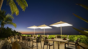 An outdoor dining area with tables and chairs, large umbrellas, and a view of the ocean at dusk.
