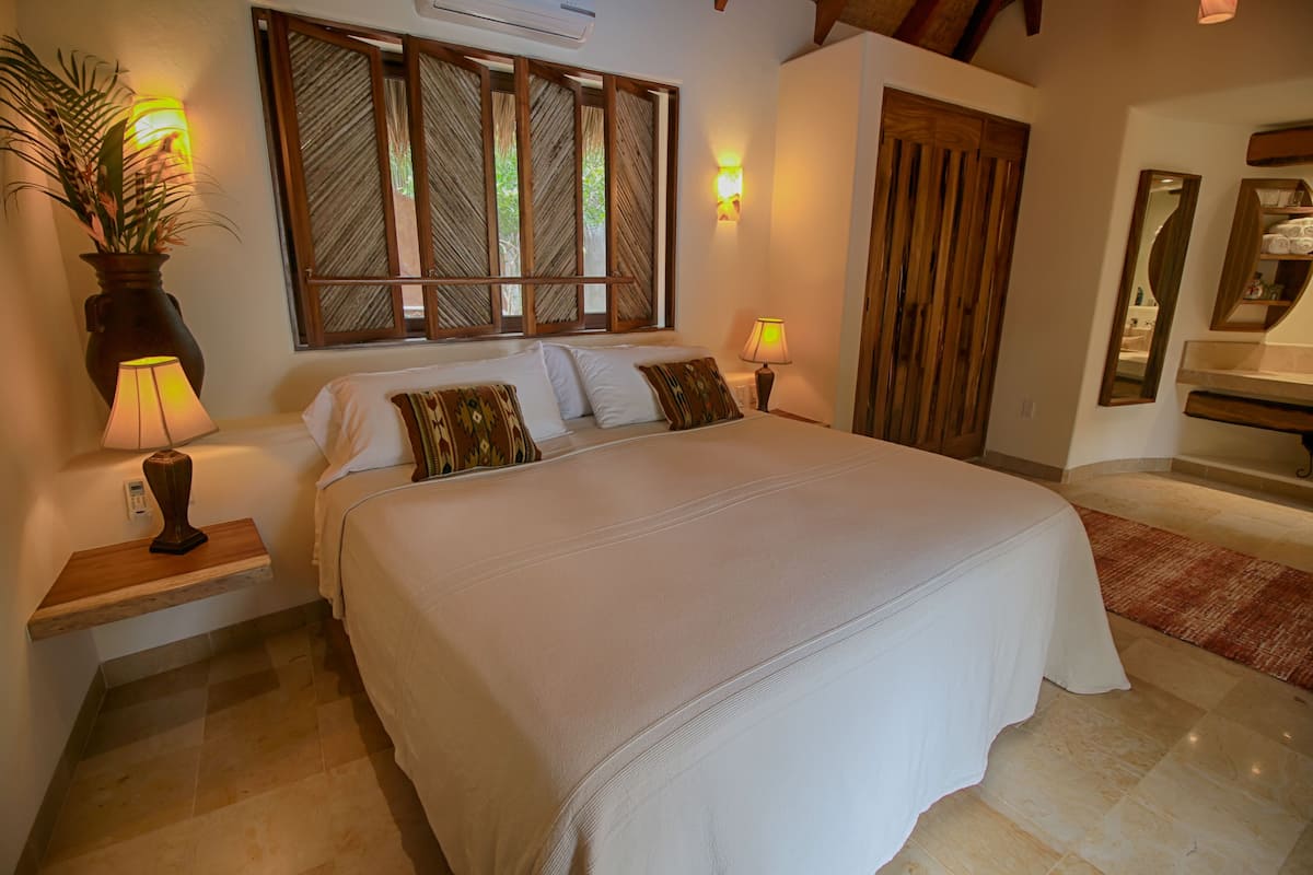 Uma cama bem arrumada com lençóis brancos e almofadas decorativas, uma mesa de cabeceira com abajur, uma planta em vaso e uma grande janela com persianas de madeira.