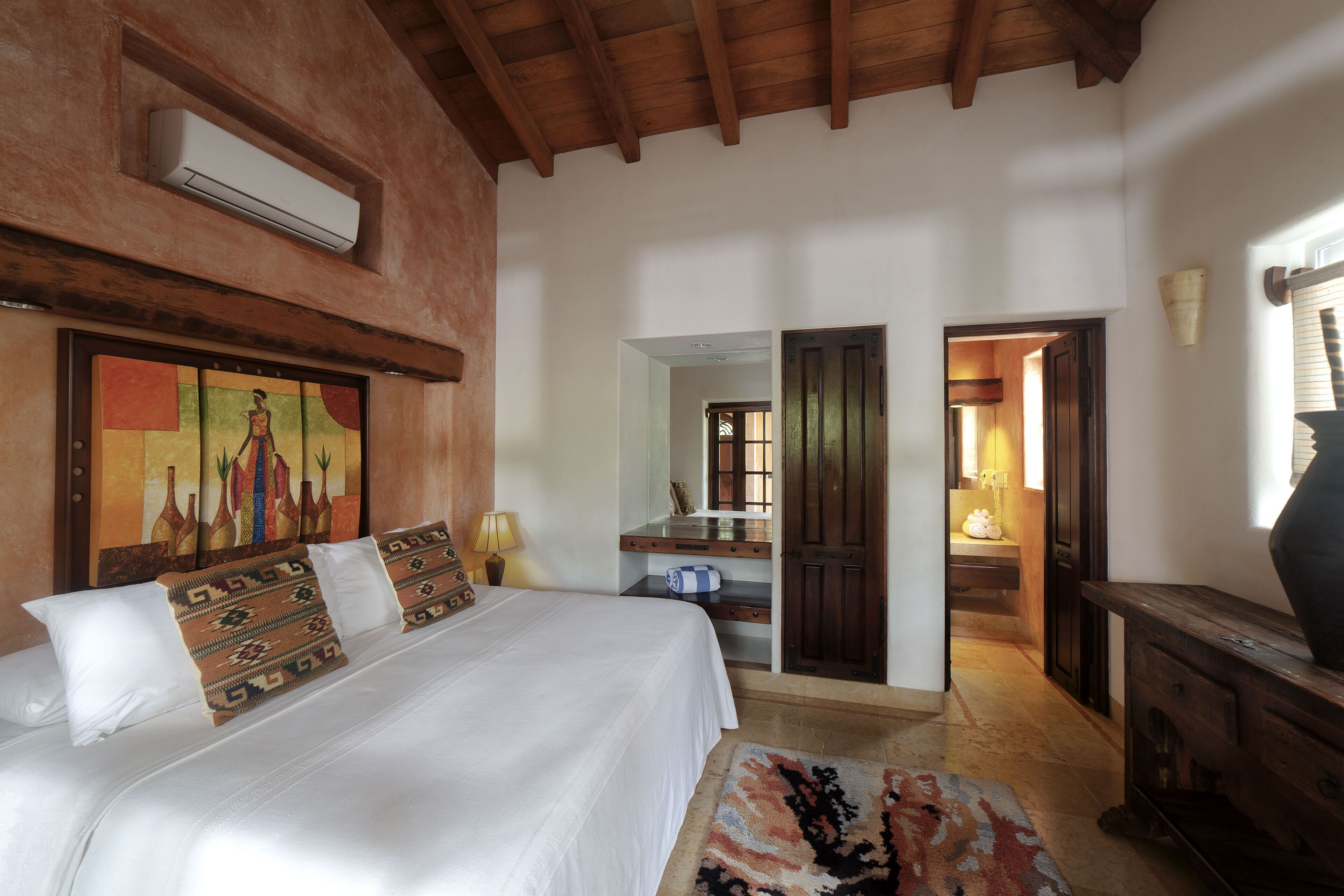 A bedroom with a large bed, a wooden ceiling, a painting on the wall, and a door leading to another room.