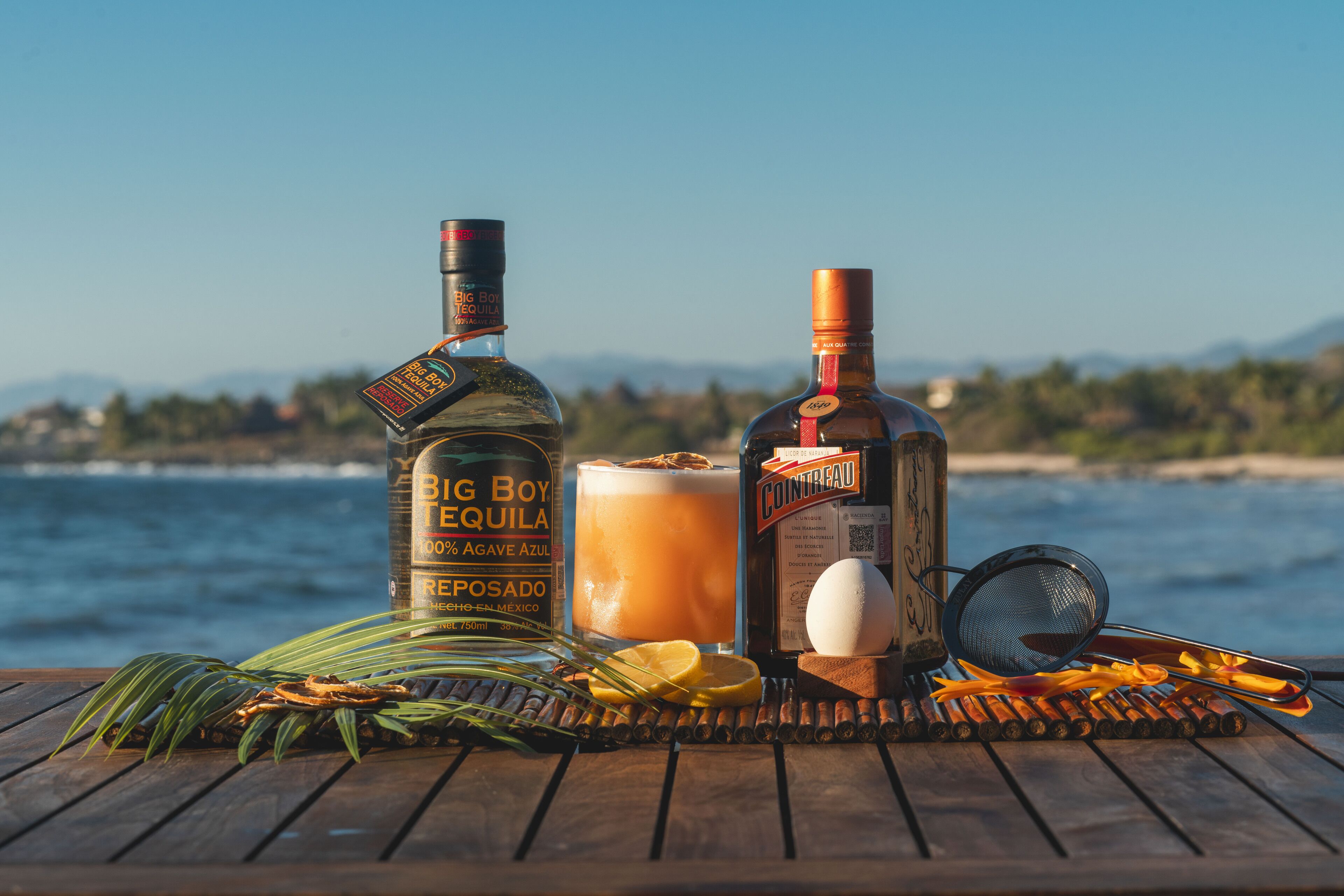 A wooden table with a bottle of Big Boy Tequila, a glass of drink, and a bottle of Cointreau, with a scenic view of the ocean and mountains in the background.