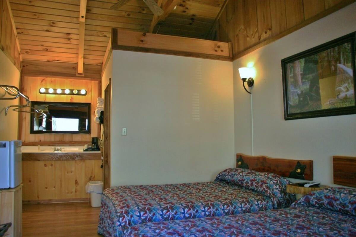 Ein gemütliches Blockhauszimmer mit Holzboden, einem Bett mit gemusterten Textilien, einer kleinen Küchenzeile mit Spüle und Spiegel sowie einem gerahmten Bild an der Wand.