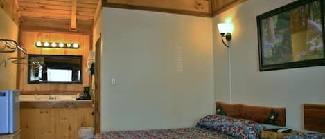 A cozy cabin-style room with a wooden ceiling, a bed with patterned bedding, a small kitchenette with a sink and a mirror, and a framed picture on the wall.