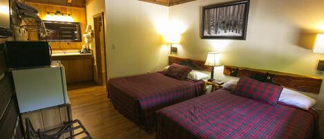 A room with two beds, a wooden ceiling, a kitchen area with a sink, and a framed picture on the wall.