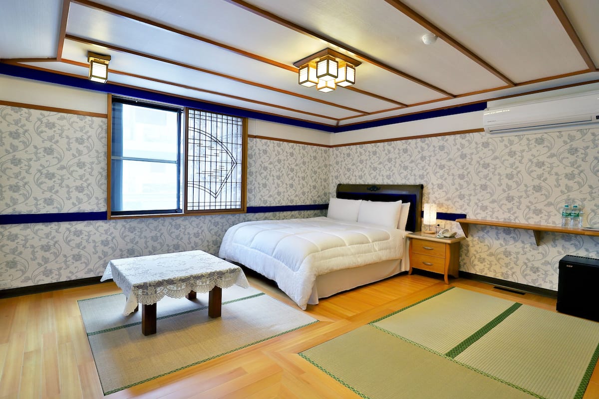 A traditional Japanese-style bedroom with tatami flooring, a futon bed, a small table with a lace tablecloth, and a window with a grid pattern.