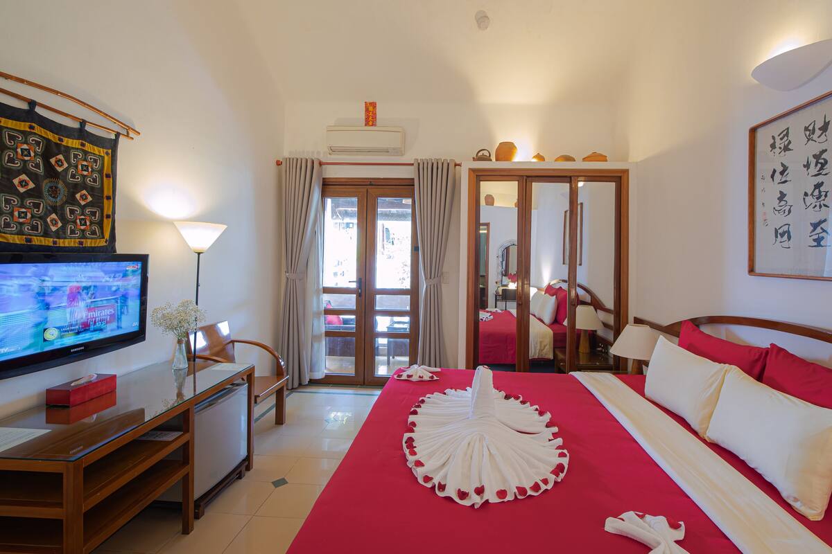 Habitación de hotel con televisión, escritorio, cama con colcha roja y almohadas blancas, espejo y un cuadro en la pared.