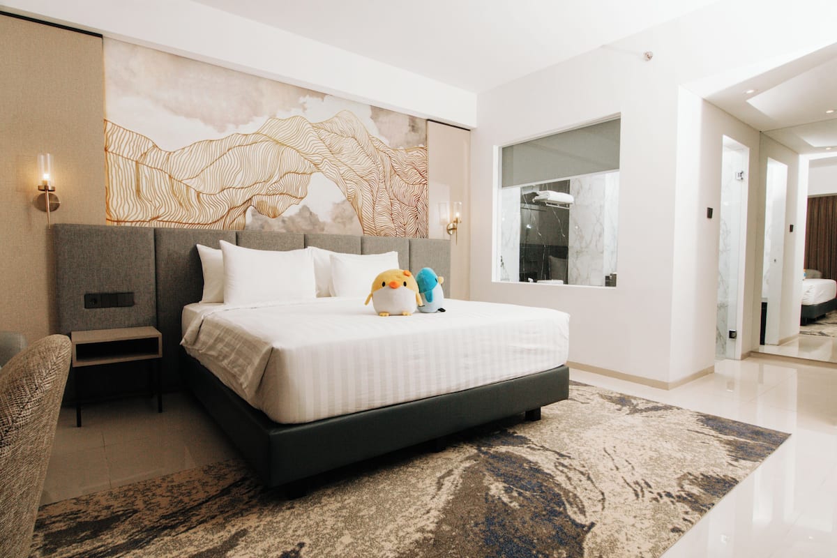 Habitación de hotel moderna con una cama grande, una mesita de noche, una lámpara de pared y un ventanal amplio.