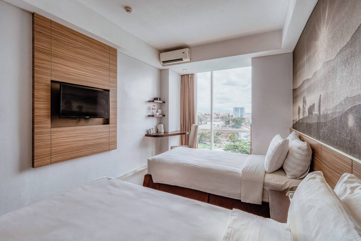Habitación de hotel con dos camas, televisor montado, equipo de aire acondicionado, ventana con vista a la ciudad y repisa con artículos de tocador.