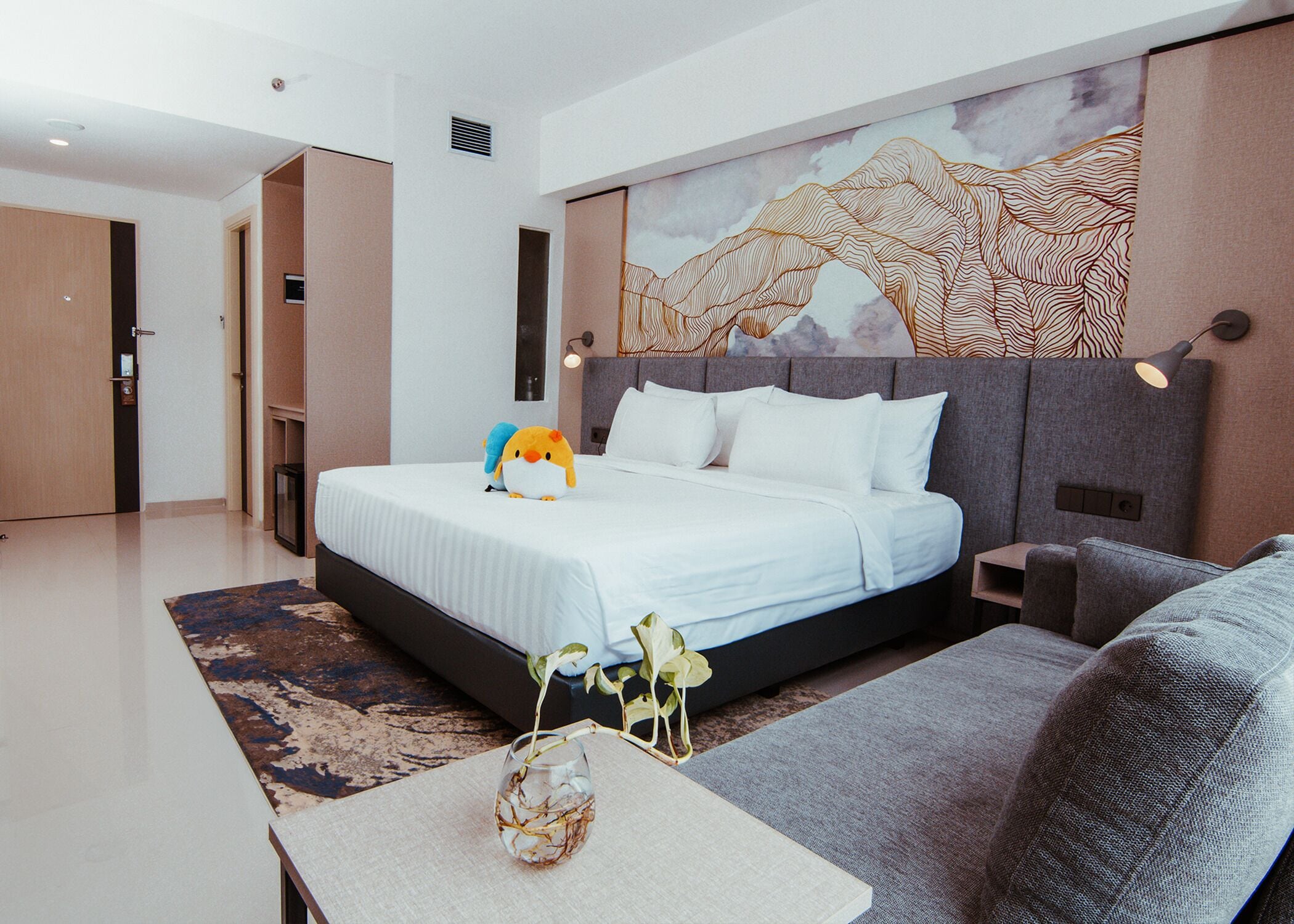 Een moderne hotelkamer met een groot bed, een bank, een kleine tafel met een vaas en een muur met een schilderij van een berglandschap.