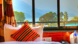 Une chambre d’hôtel avec un lit, une fenêtre donnant sur les montagnes et un lac, une bouteille d’eau et un téléphone sur la table de chevet.