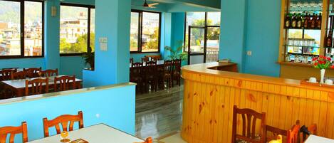 A restaurant with blue walls, wooden tables and chairs, a bar area, and large windows offering a view of buildings and trees.