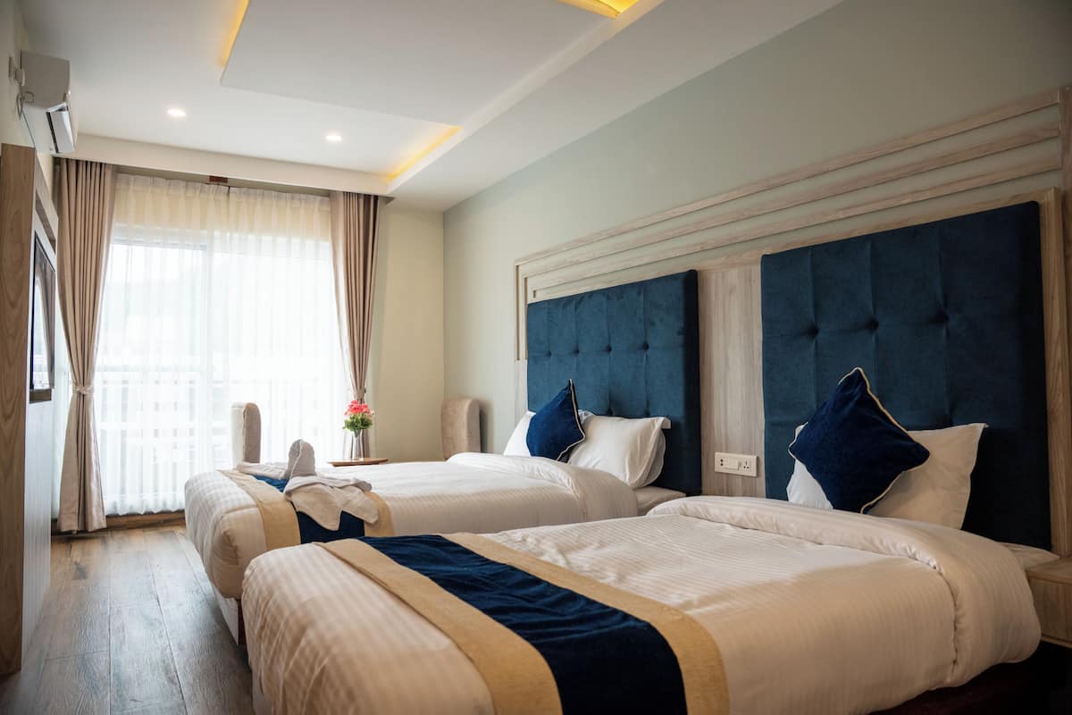 Ein Hotelzimmer mit zwei Betten, einem großen Fenster mit Vorhängen, einem Nachttisch mit Blumen und einer Wand-Klimaanlage.