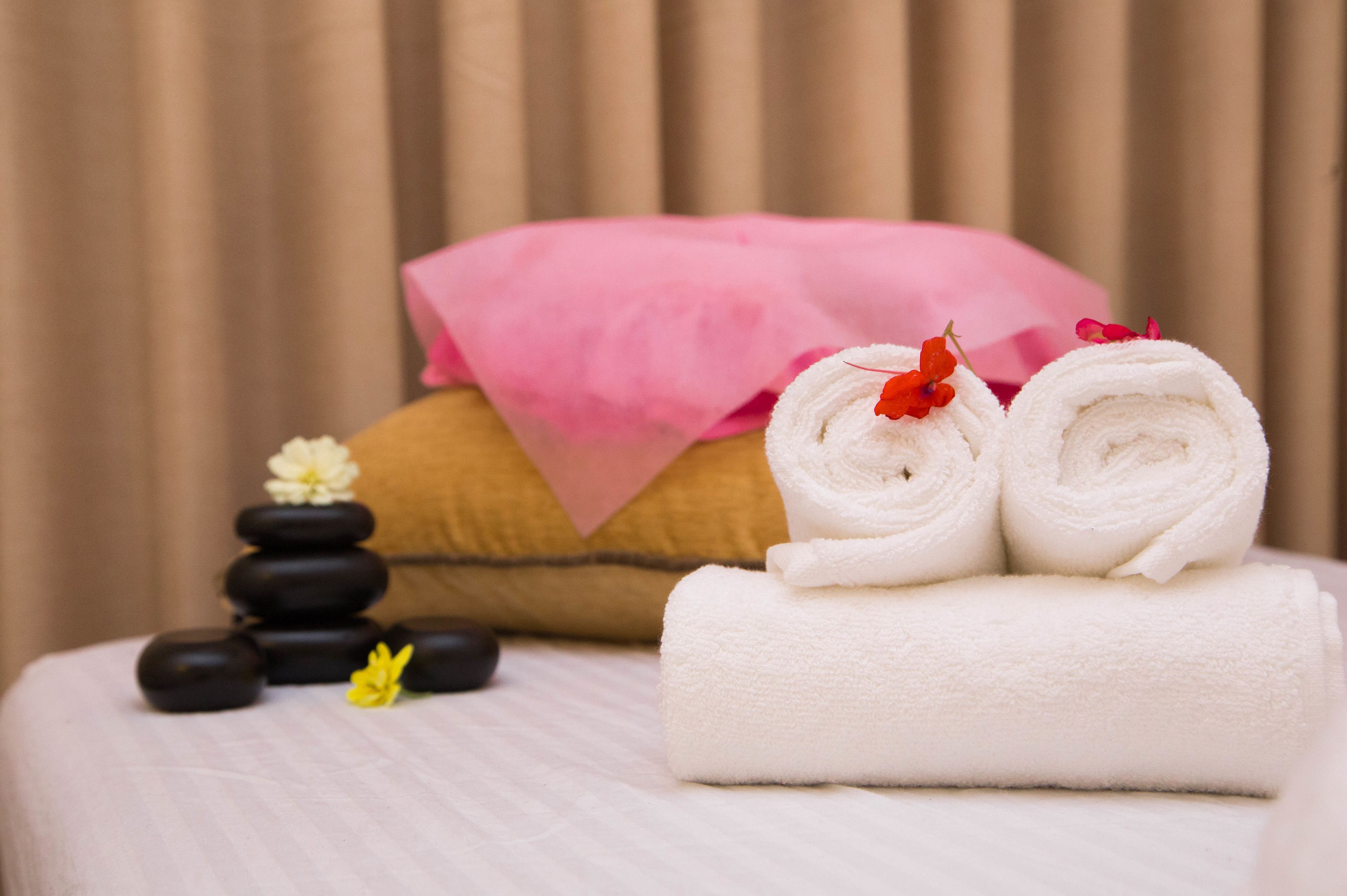 A neatly arranged massage area with a bed, rolled towels, a pink towel, and a stack of black stones.