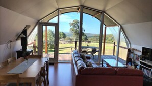 A cozy living area with a sofa, a dining table, and a bookshelf. A large window offers a view of a grassy field and trees. - Tennessee Hill Chalets (Lowlands)