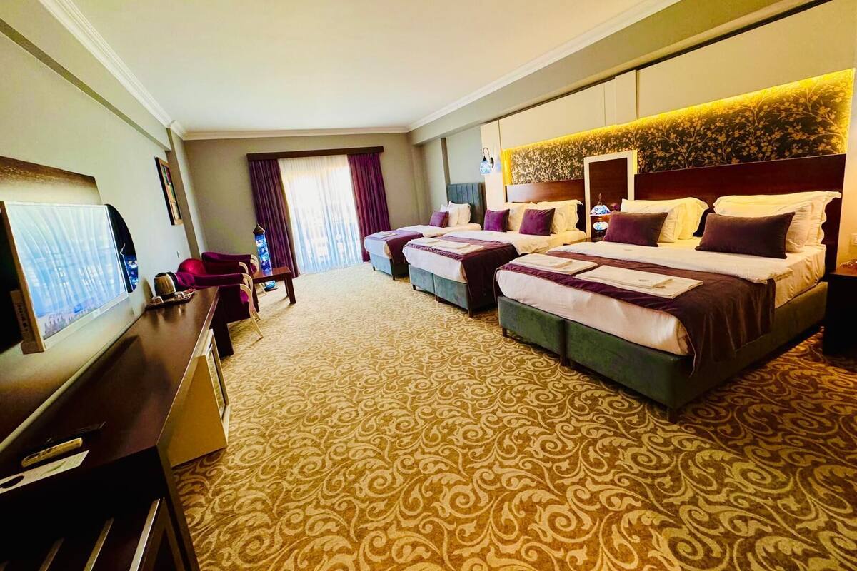 Ein Hotelzimmer mit einem großen Bett, einem kleineren Bett, einem Schreibtisch, einem Fernseher und einem Fenster mit Vorhängen.