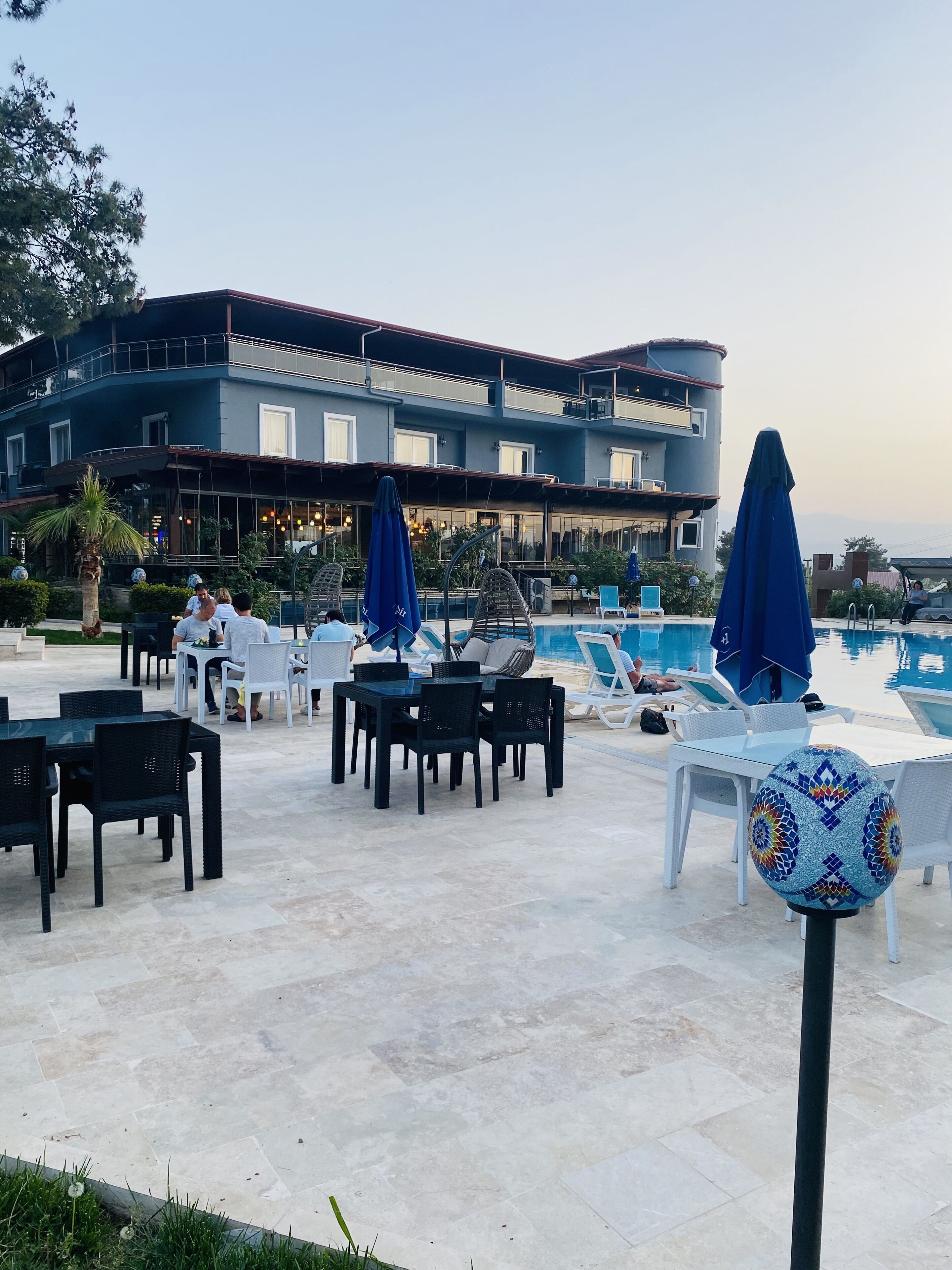 A poolside area with lounge chairs and umbrellas, a dining area with tables and chairs, and a building with large windows and a balcony.