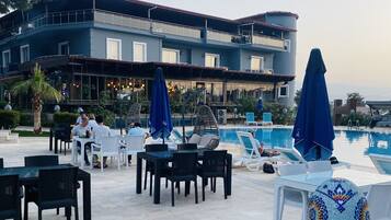 A poolside area with lounge chairs and umbrellas, a dining area with tables and chairs, and a building with large windows and a balcony.