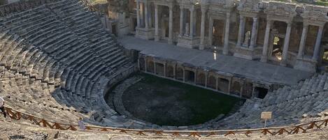 Un antiguo anfiteatro con asientos escalonados y un escenario central, rodeado de ruinas y campos abiertos.
